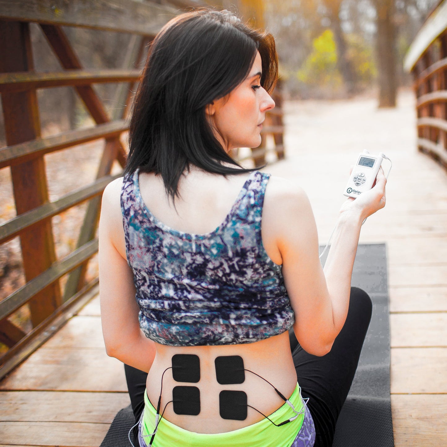 Woman wearing iReliev TENS electrode pads on lower back, holding wireless remote control device outdoors.