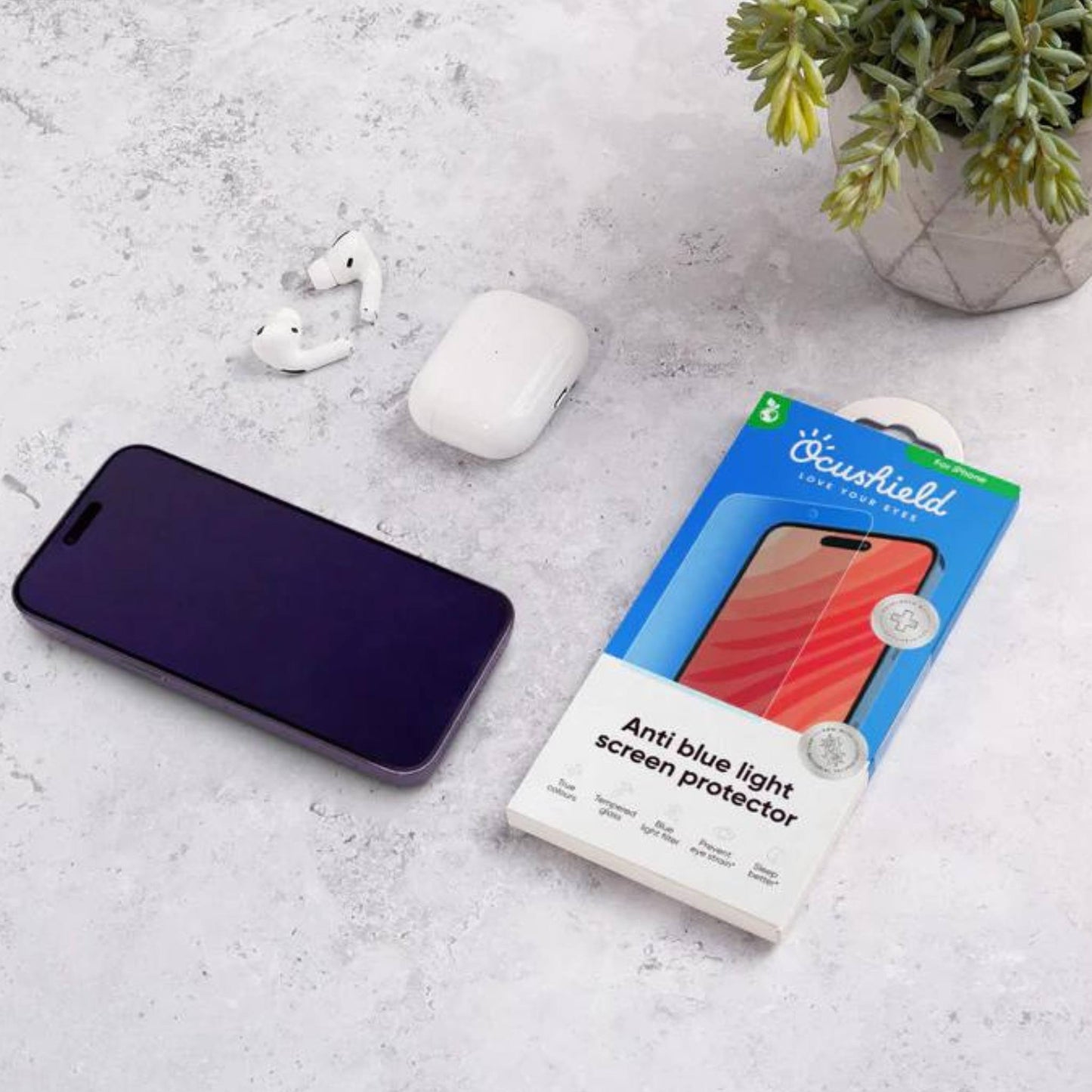 Ocushield blue light screen protector packaging displayed next to smartphone, earbuds, and potted plant on concrete surface.