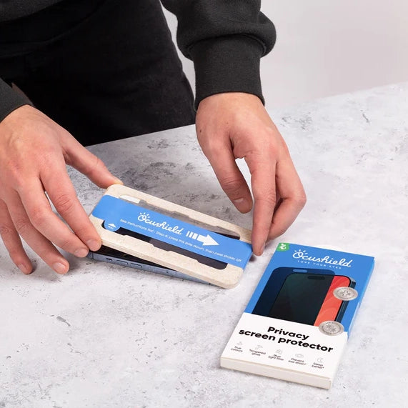 Hands applying blue Ocushield Privacy Screen Protector to iPhone next to product packaging on marble surface.