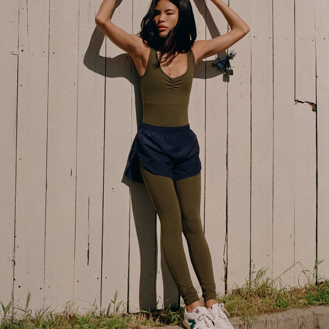 Juunaday Even Flow Unitard in olive with navy shorts, styled with white sneakers against a white wooden wall.