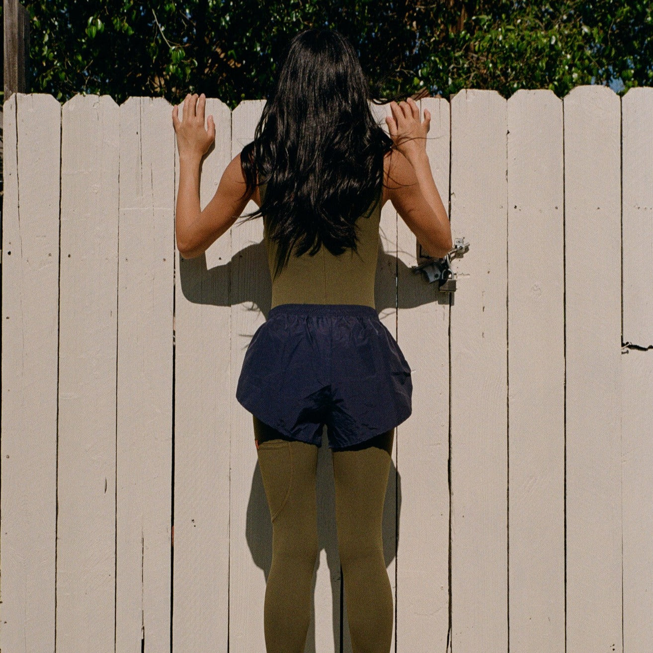 Woman wearing Juunaday Even Flow Unitard in olive and navy, rear view against white fence.