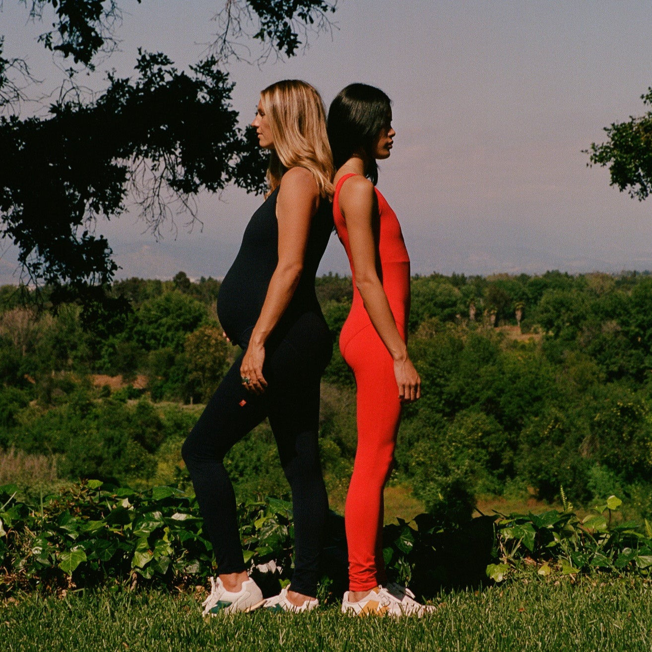 Two women wearing Juunaday Even Flow Unitards in black and red standing back-to-back outdoors with forest landscape background.