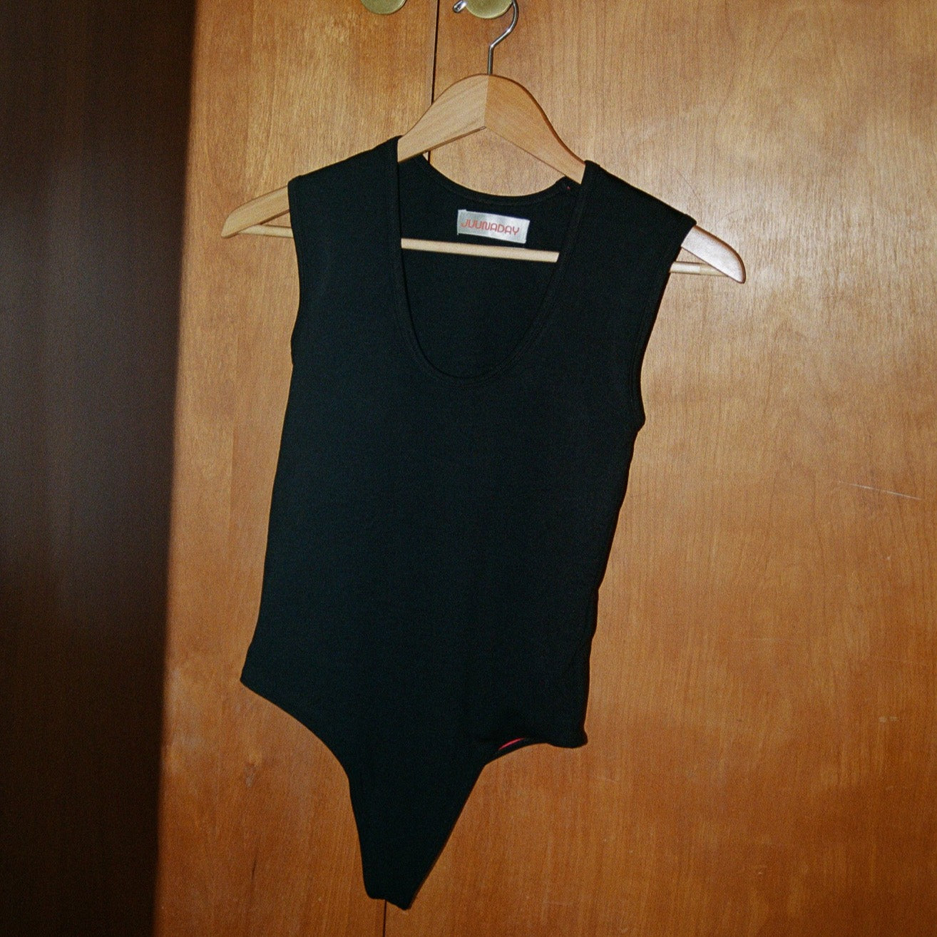 Black Juunaday All Day Bodysuit displayed on wooden hanger against wood paneled wall, front view.