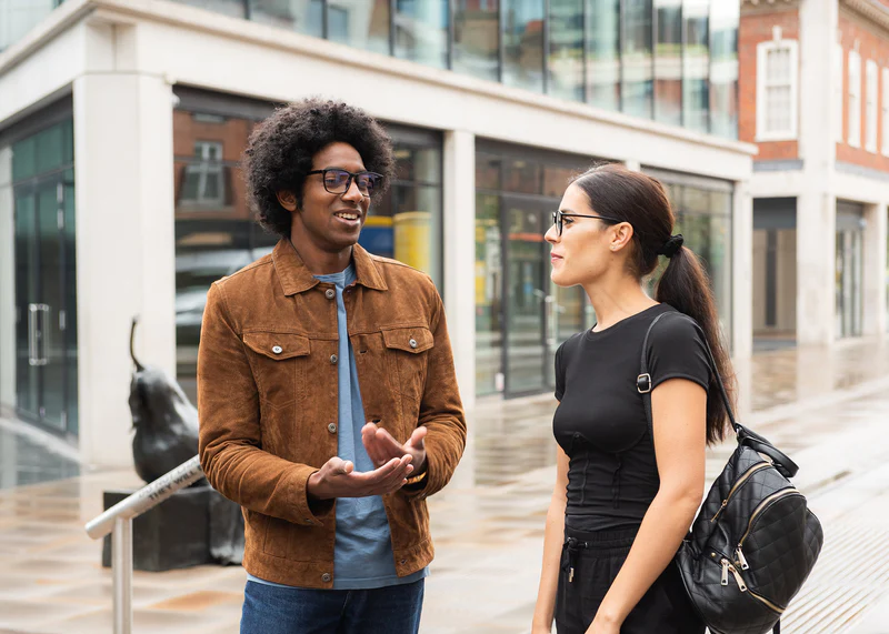 Two people wearing glasses converse outdoors on a modern urban street, one in Ocushield Clear blue light glasses with a brown jacket.