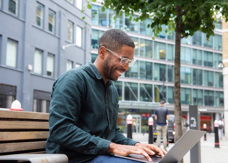 Man wearing Ocushield Clear blue light glasses while working on laptop outdoors on city bench.