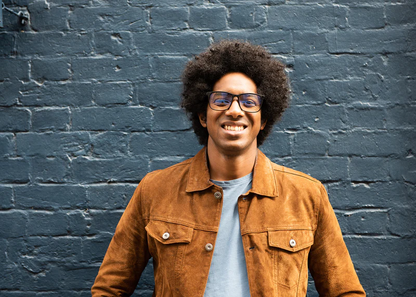 Man wearing Ocushield Clear blue light glasses and tan jacket smiling against dark blue brick wall.