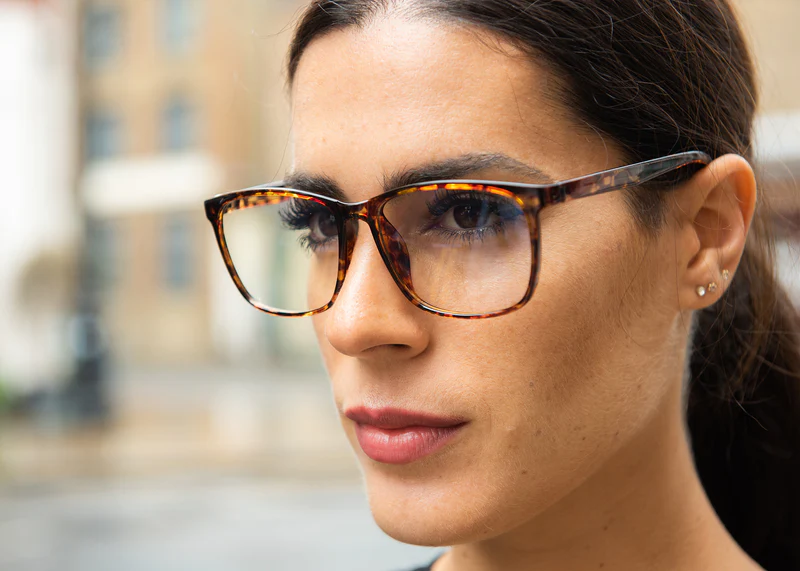 Woman wearing tortoiseshell Ocushield Clear blue light glasses, side profile view, in outdoor setting.