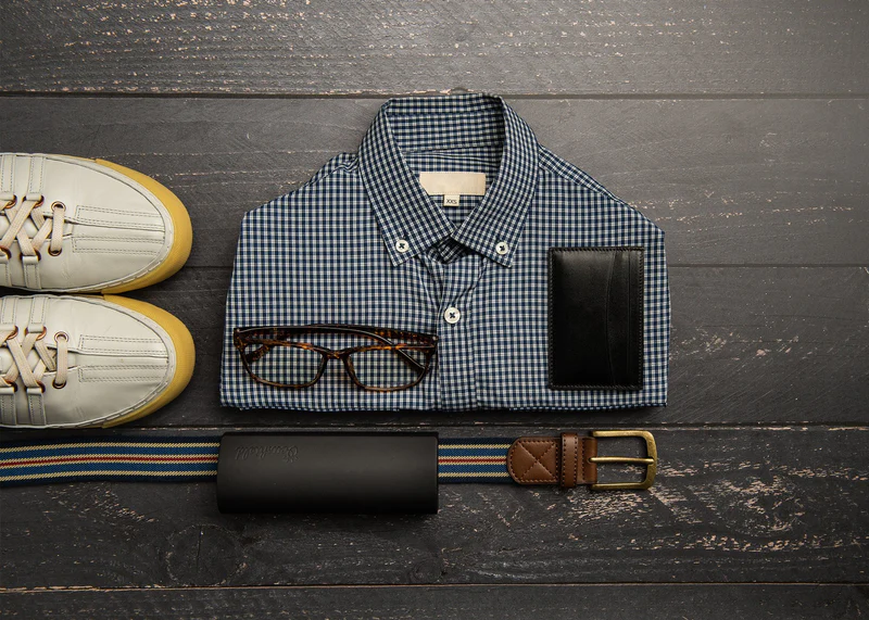 Ocushield Clear blue light glasses displayed on folded checkered shirt with shoes, belt, wallet, and phone in flat lay arrangement.