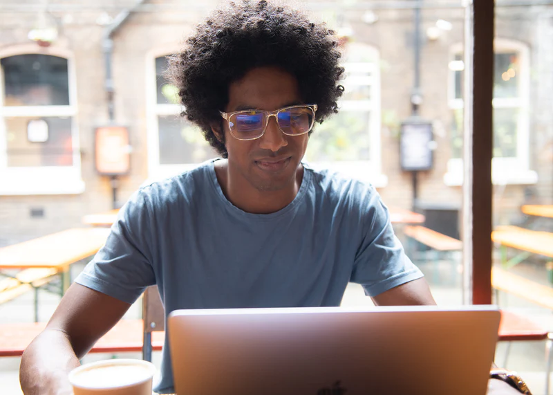 Young man wearing Ocushield Clear blue light glasses while working on laptop at bright café table.
