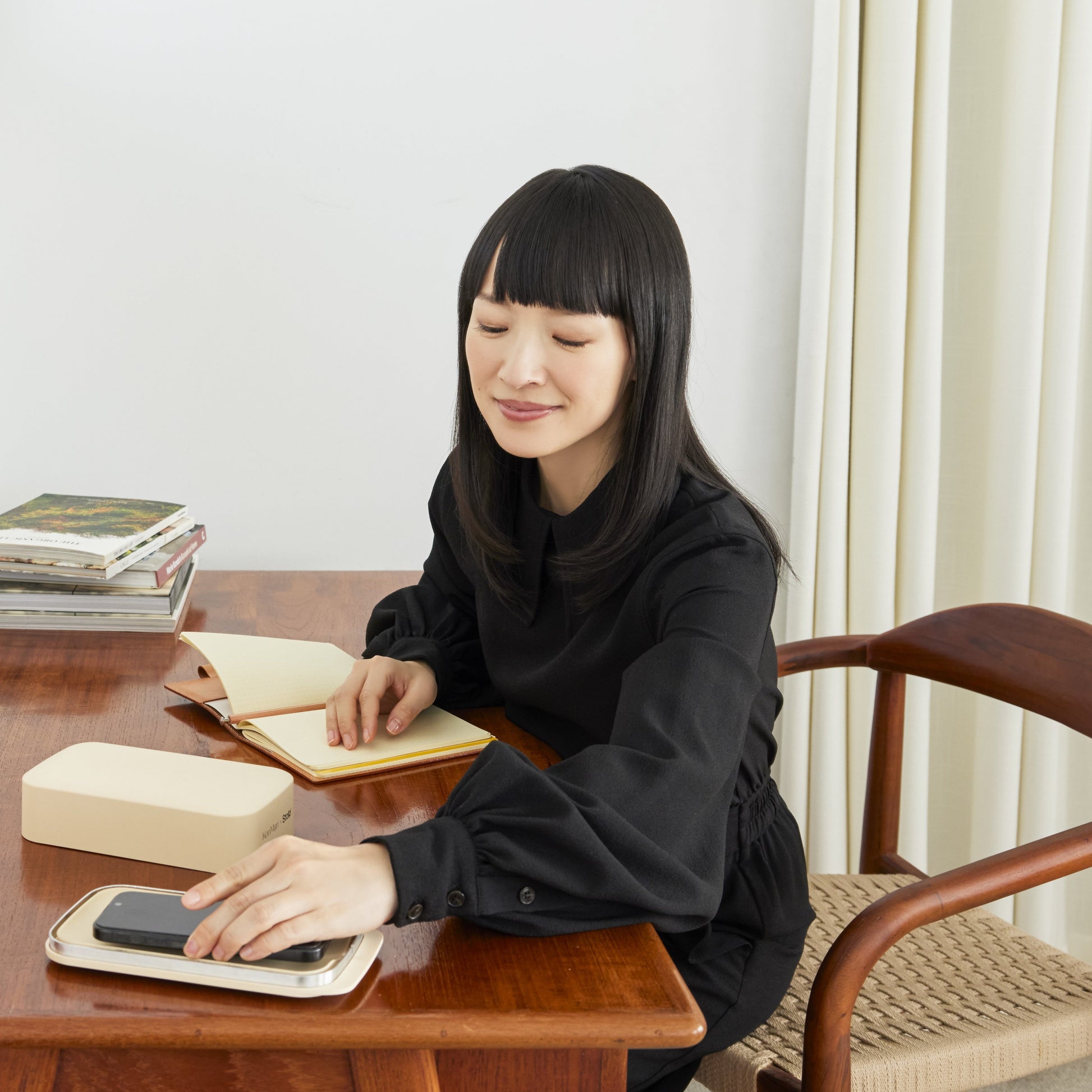 Marie Kondo seated at a wooden desk with one hand in a notebook and the other resting on a phone inside an open sand-colored tray.