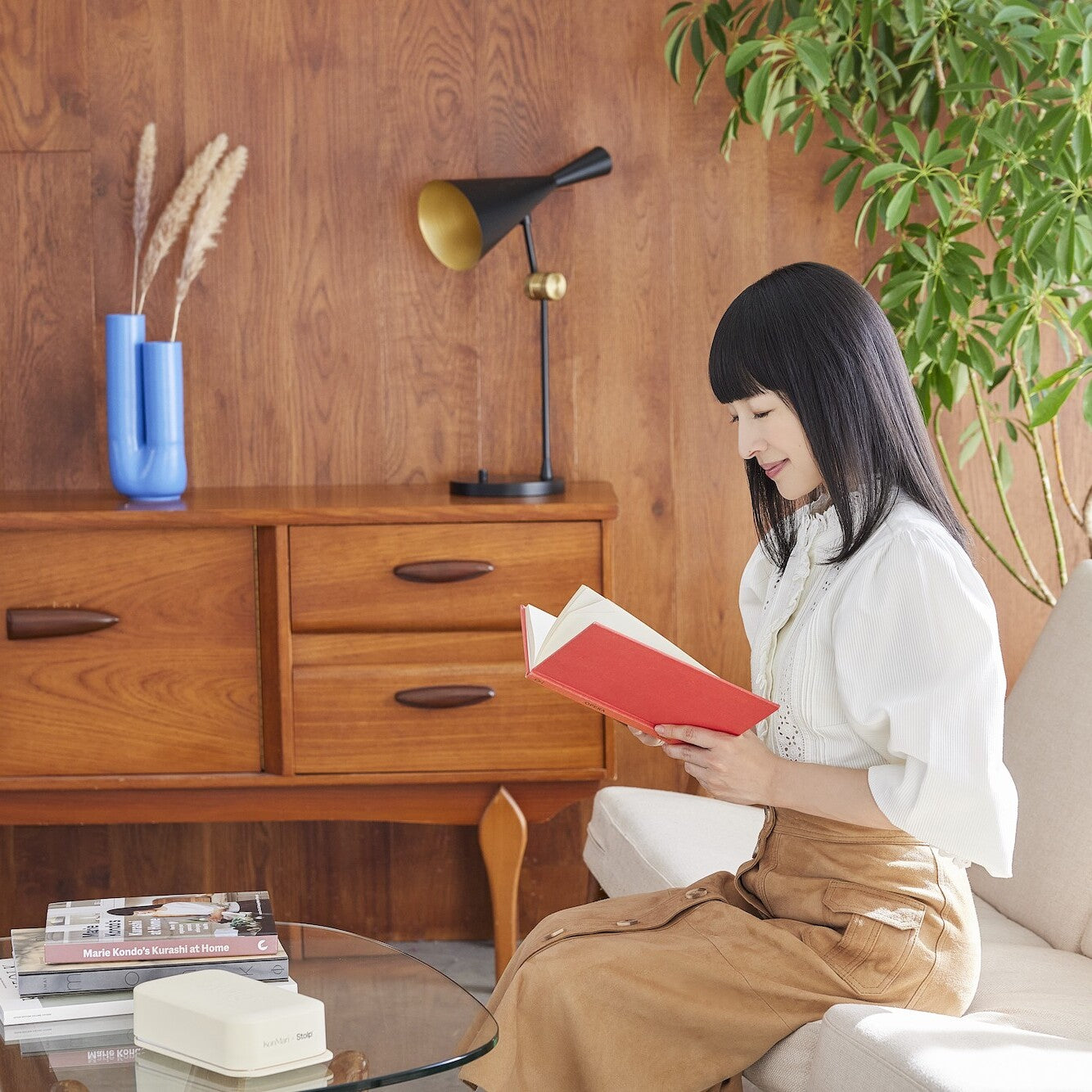 Woman reading a hardcover book on a sofa beside a sand-colored Bagby Stolp Faraday box and a stack of books on a glass table.