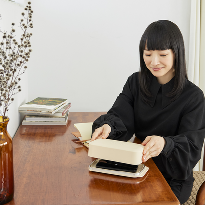 Woman at a wooden desk lifting the lid from a sand-colored Bagby Stolp Faraday box, revealing a phone resting in the tray.
