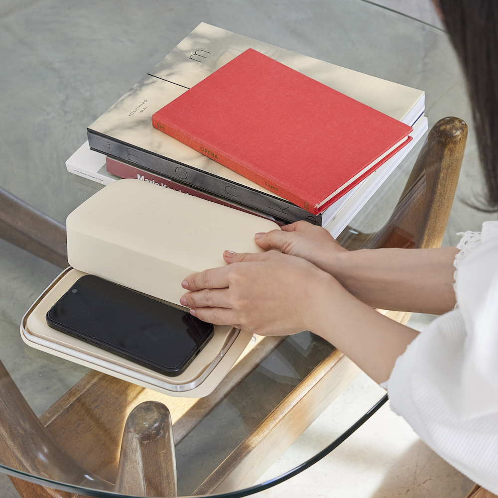 Overhead view of a phone resting in an open sand-colored Bagby Stolp Faraday tray on a glass table beside a stack of books.