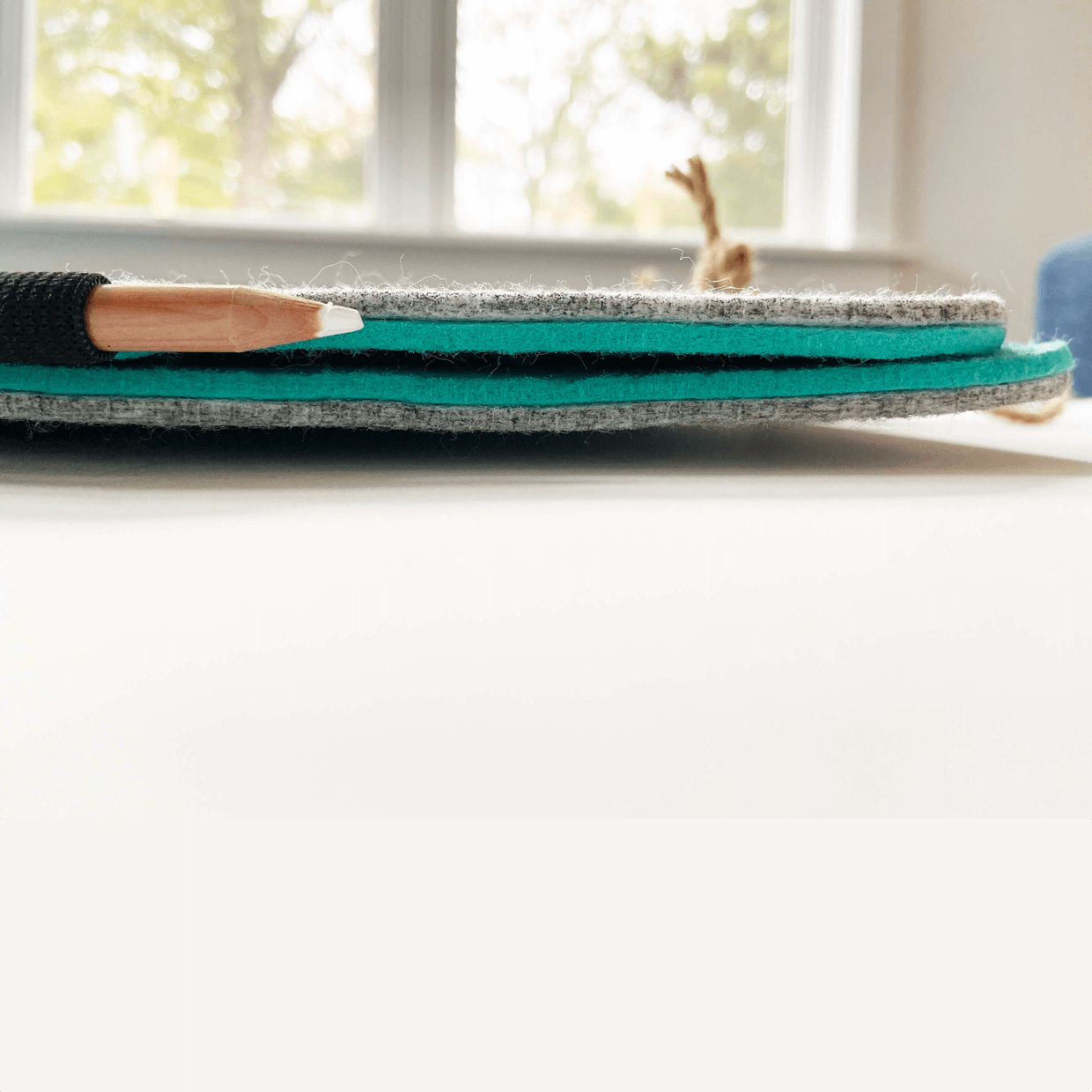 Bagby 4 Pocket Phone Holder displayed flat, showing layered gray and teal felt construction with a paintbrush resting on top near a bright window.