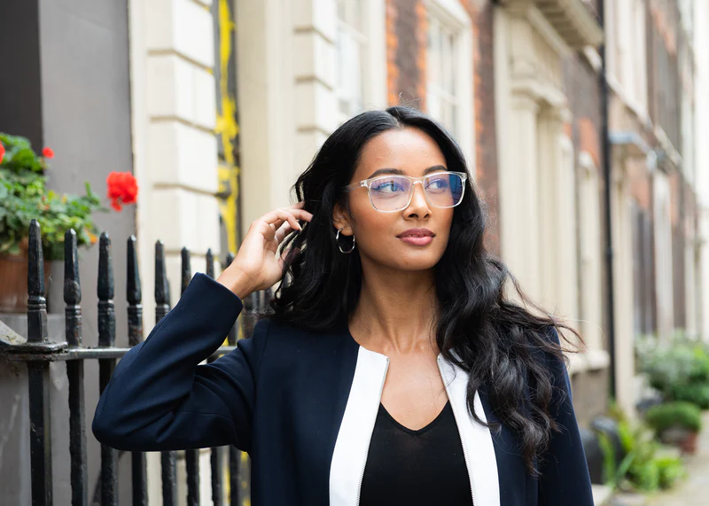 Woman wearing Ocusheel Clear blue light glasses with clear frames, navy blazer, and black top, standing on urban street.