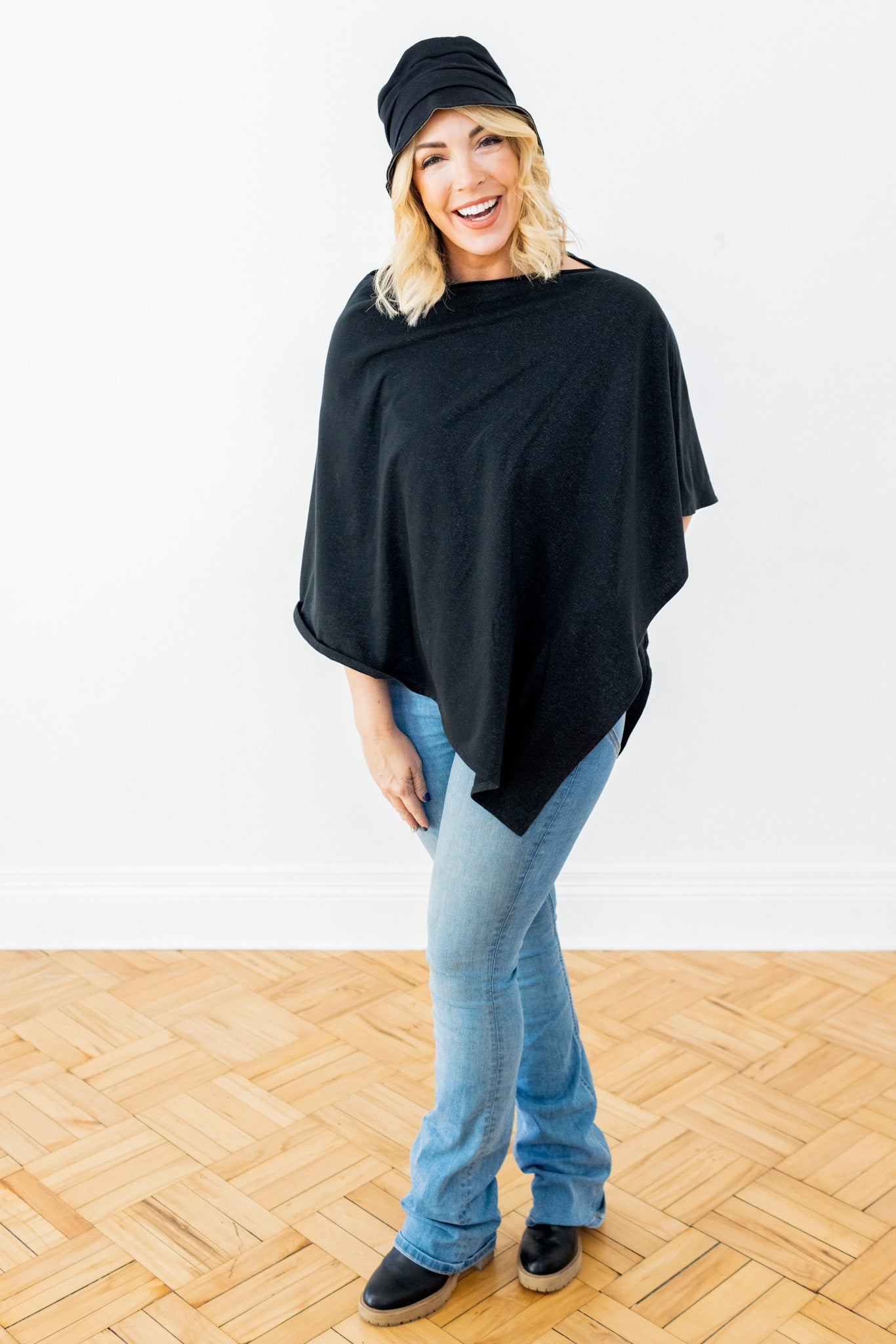 Woman wearing black bucket hat and black poncho over light blue jeans, smiling at camera on wooden floor.