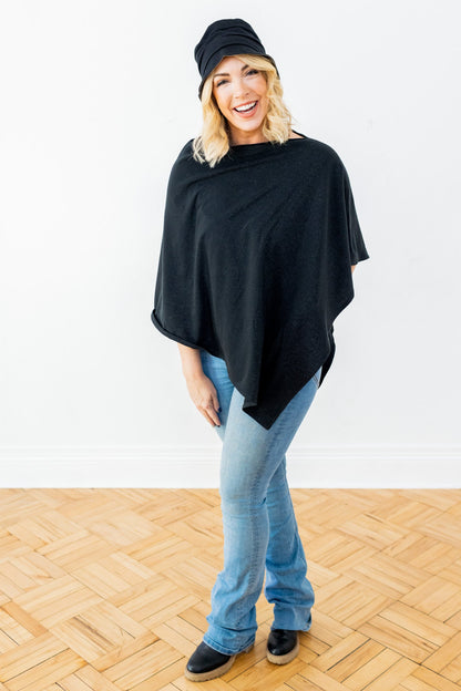 Woman wearing black bucket hat and black poncho over light blue jeans, smiling at camera on wooden floor.