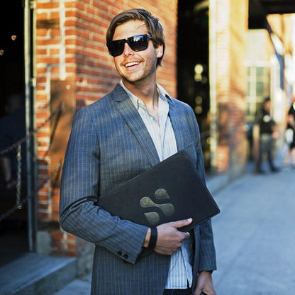 Man in pinstriped blazer holding black SafeSleeve laptop case while wearing sunglasses on urban street.