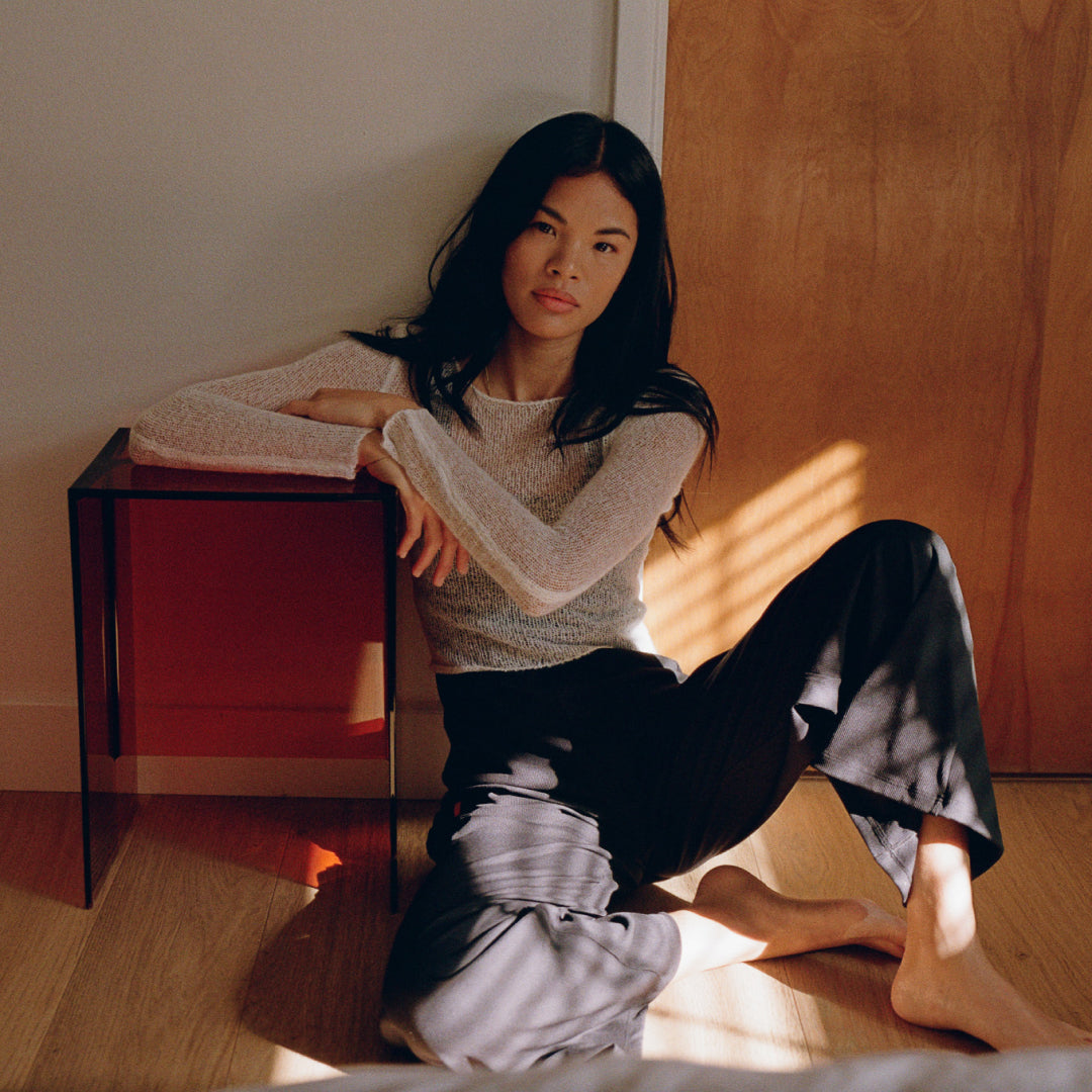 Woman sitting on wooden floor wearing beige knit sweater and black Juunaday Everywhere Playsuit with wide legs, sunlight streaming across scene.