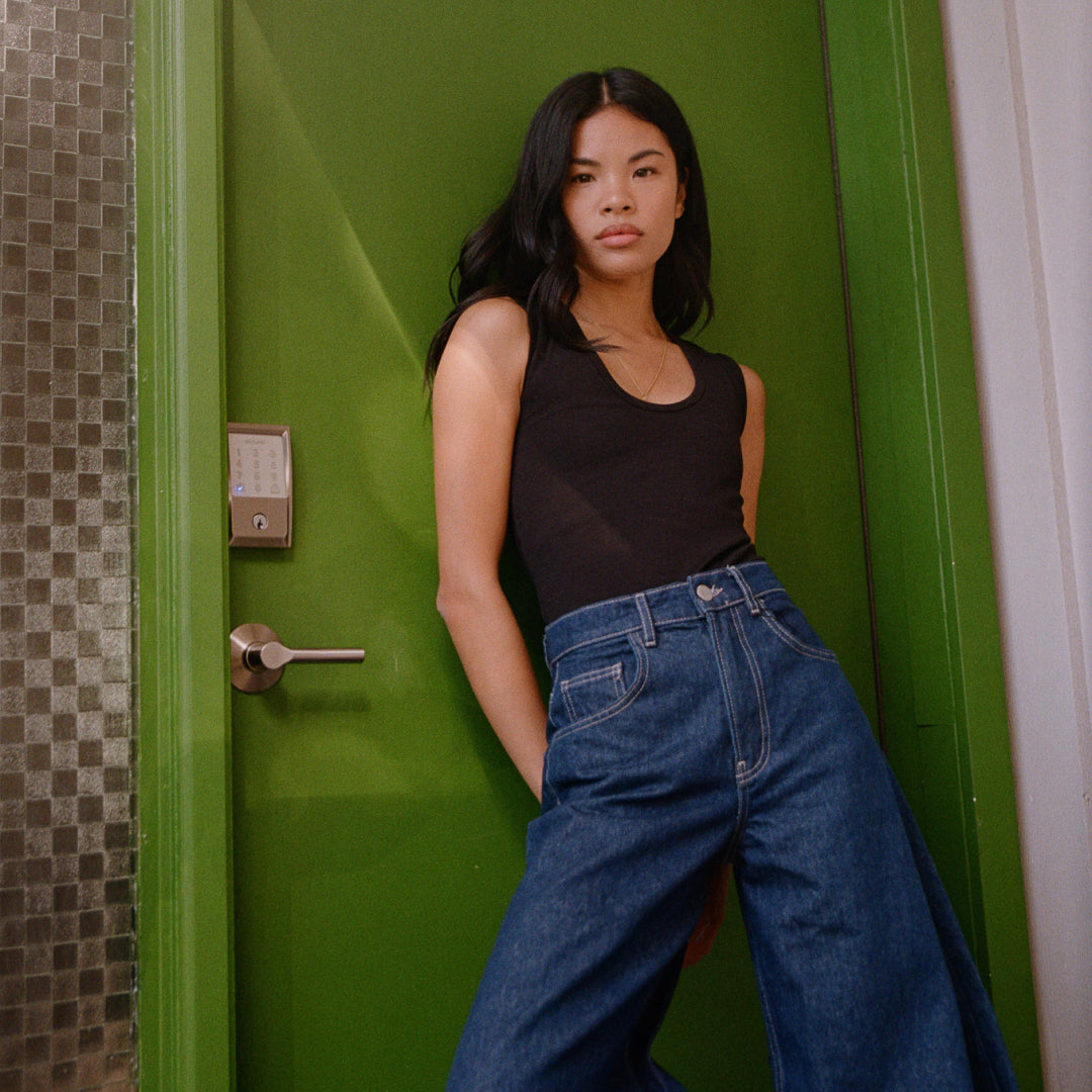Woman wearing black Juunaday All Day Bodysuit with denim jeans, leaning against bright green door in hallway setting.