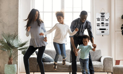 A family of four dancing together in a bright living room, with a gray Bagby FAMILY organizer hanging on the wall behind them.