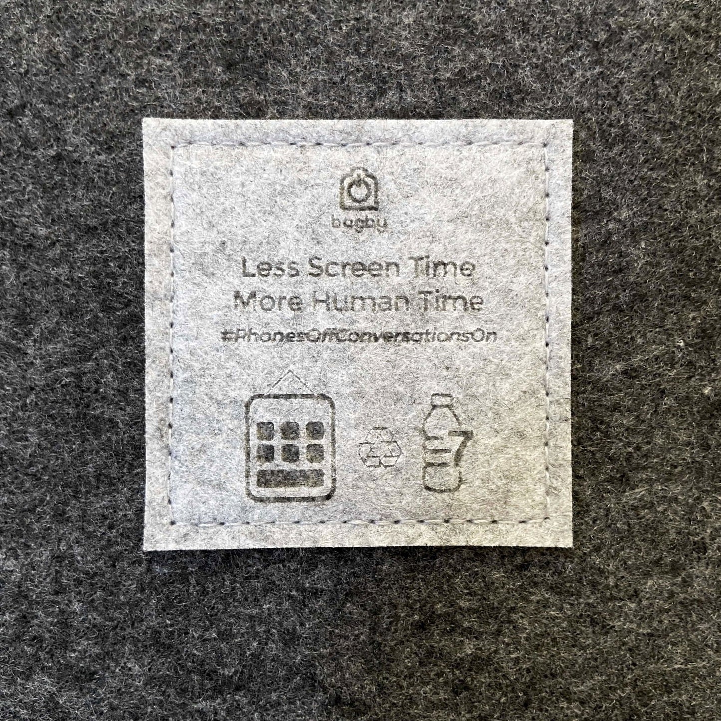 Close-up of the embroidered Bagby label reading "Less Screen Time, More Human Time" with recycling and phone icons on dark gray felt.