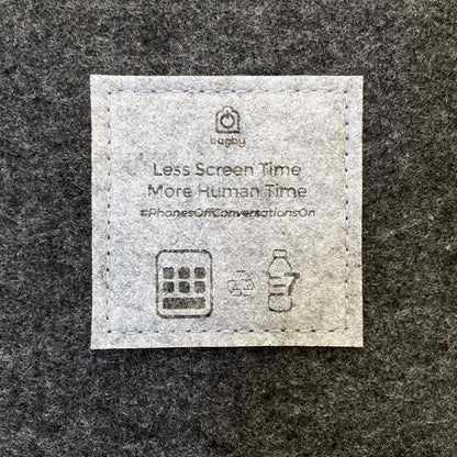 Close-up of the embroidered Bagby label reading "Less Screen Time, More Human Time" with recycling and phone icons on dark gray felt.