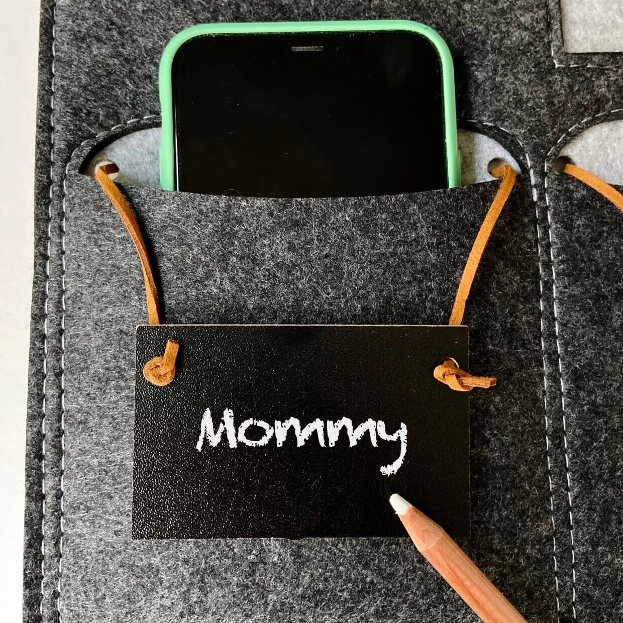 Close-up of a green-cased smartphone tucked into a dark gray felt pocket above a chalkboard tag handwritten "Mommy".