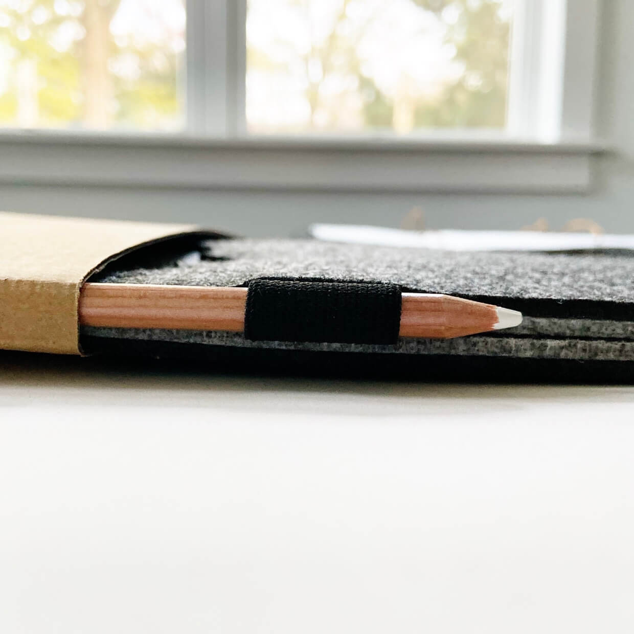 Close-up of a wooden chalk pencil held in a black elastic loop on the side of the dark gray felt organizer, backlit by a window.