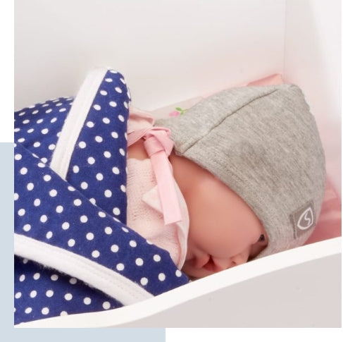 Newborn baby wearing gray SYB Baby Beanie with pink bow, wrapped in navy polka dot blanket in crib.