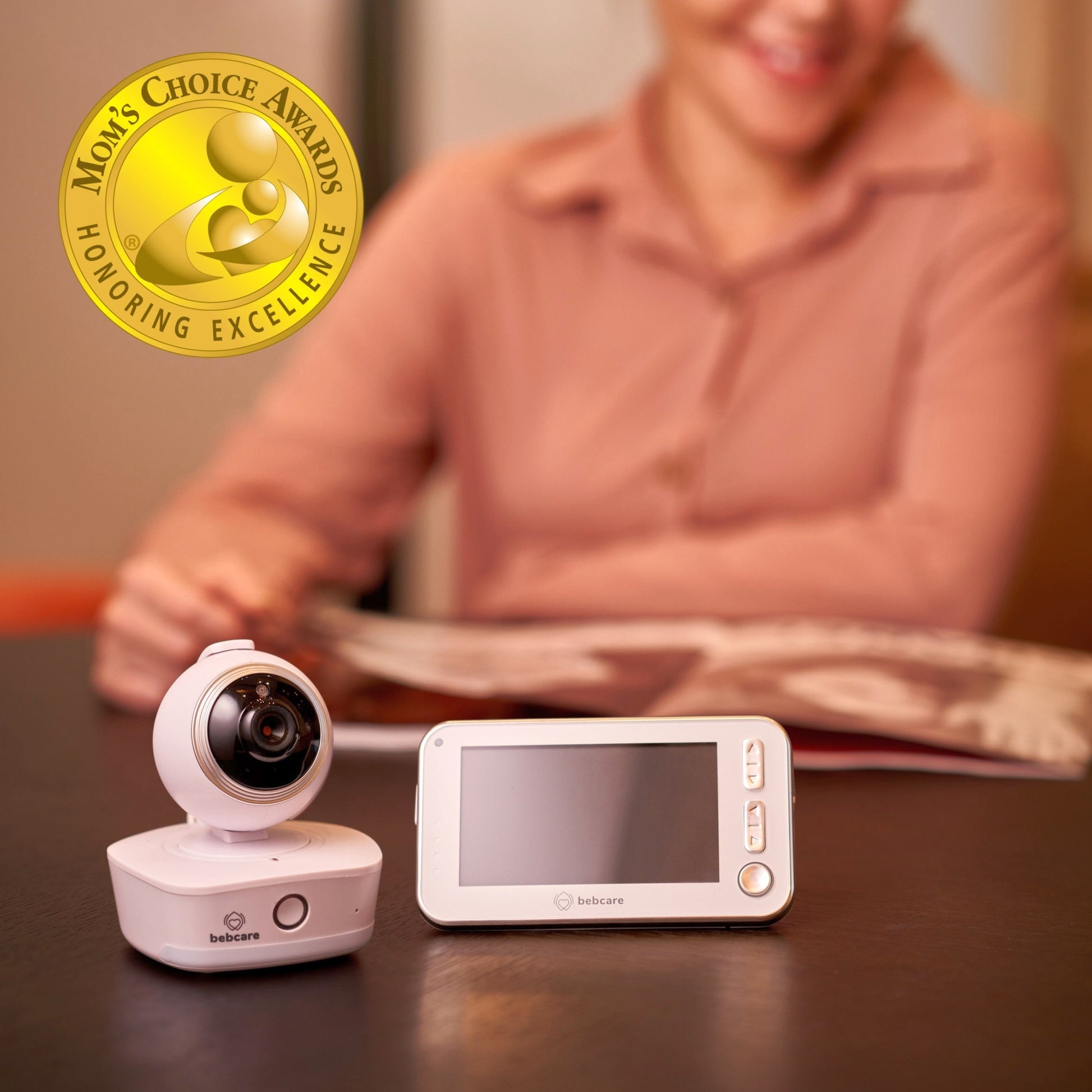 Bebcare baby monitor camera and parent unit displayed on table with Moms Choice Award seal, caregiver in background.