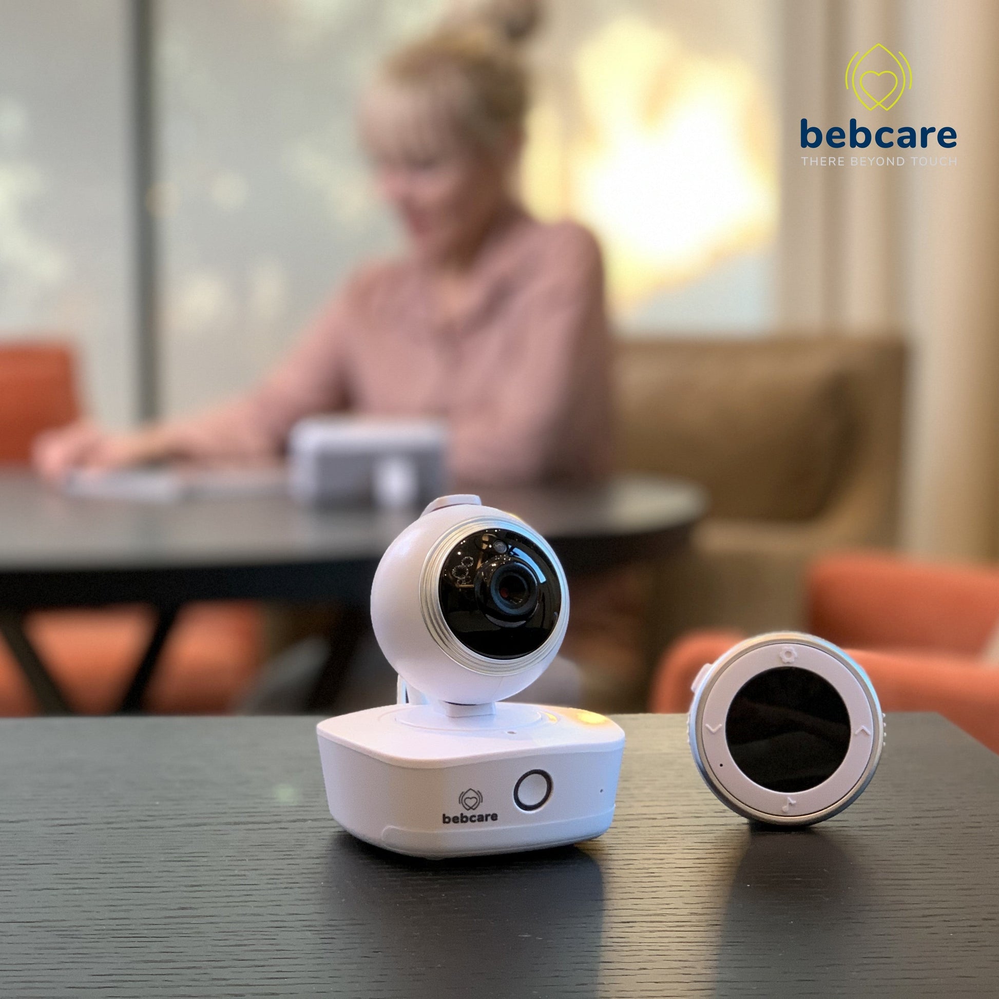 Bebcare baby monitor camera displayed on table with lens cap beside it, blurred caregiver in background.