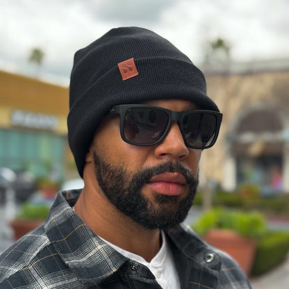 Man wearing black SafeSleeve Beanie with red patch logo and dark sunglasses, outdoor setting.