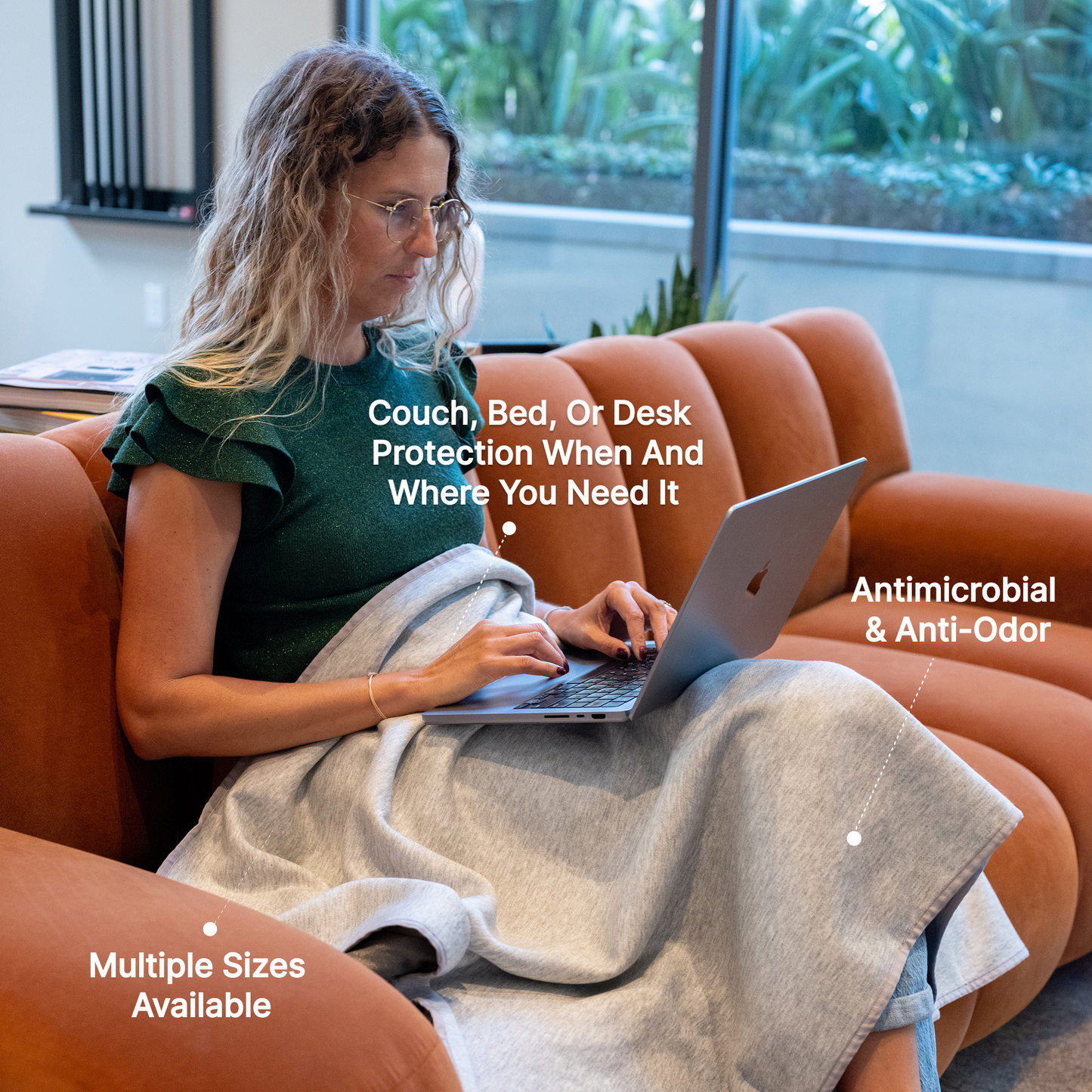 Woman using laptop on orange couch with HAVN WaveStopper™ Blanket draped across lap, highlighting antimicrobial, anti-odor features and multiple size options.