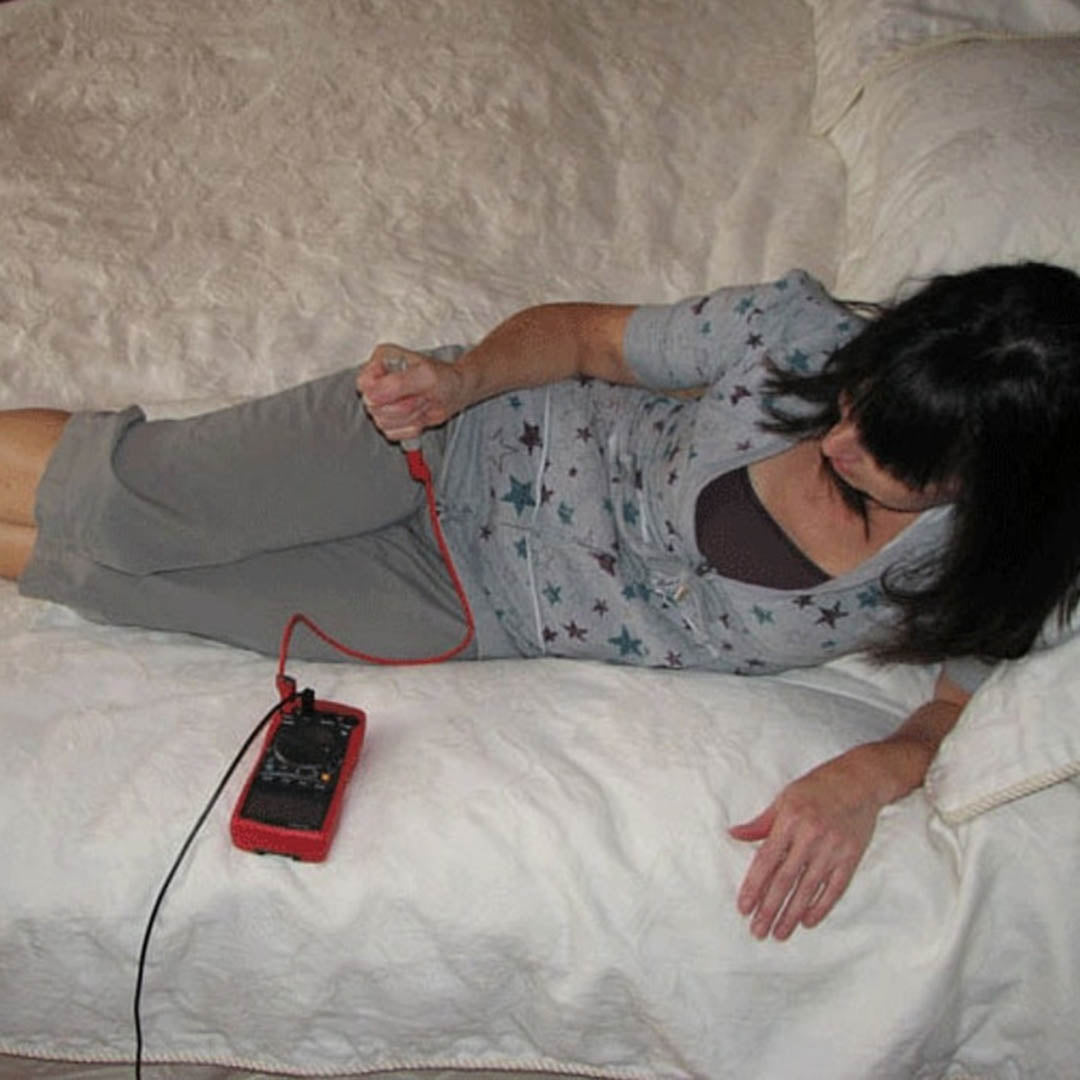 Person lying on bed using SLT Safe & Sound Body Voltage Home Test Kit with red multimeter device connected by cord.