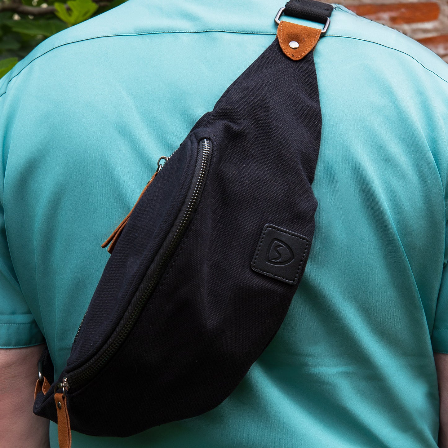 Black canvas SYB Cross Body Fanny Pack worn across chest on person in turquoise shirt, showing front zippered compartment and tan leather accents.