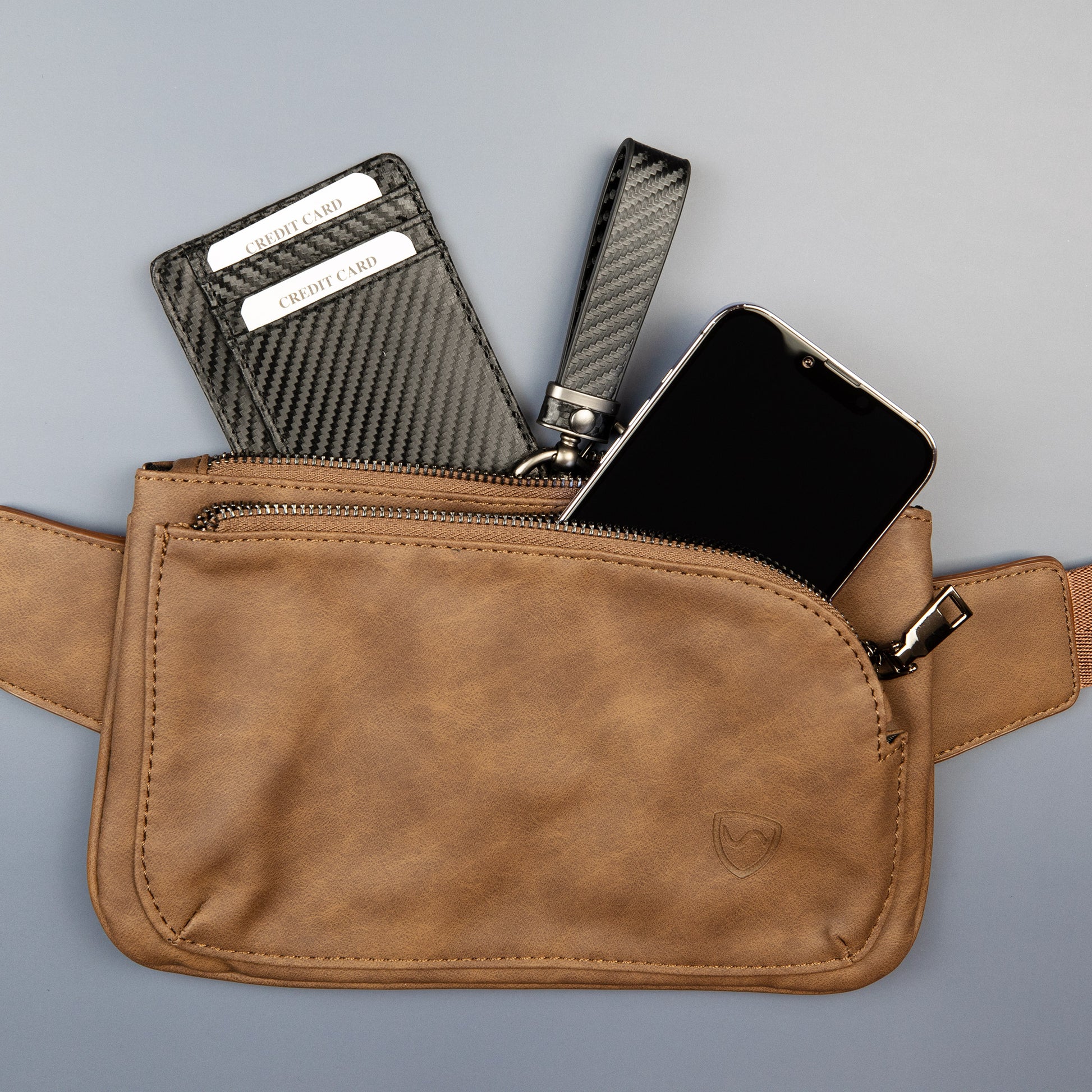 SYB Cross Body Fanny Pack in tan vegan leather displayed open, showing interior contents including a carbon fiber wallet, black comb, and smartphone.