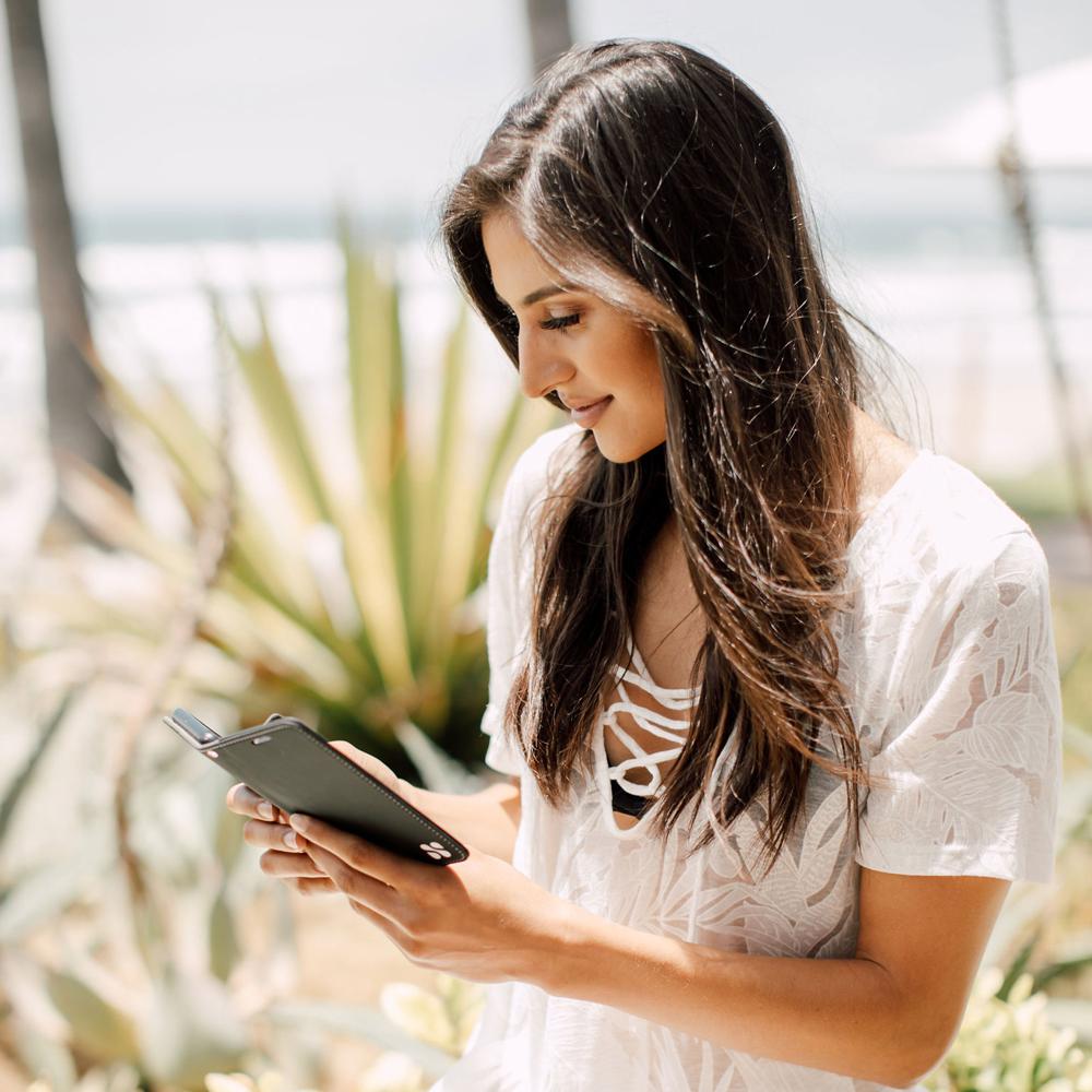 Woman in white shirt holding Samsung Galaxy phone with SafeSleeve Detachable Case, ocean view and palm plant in background.