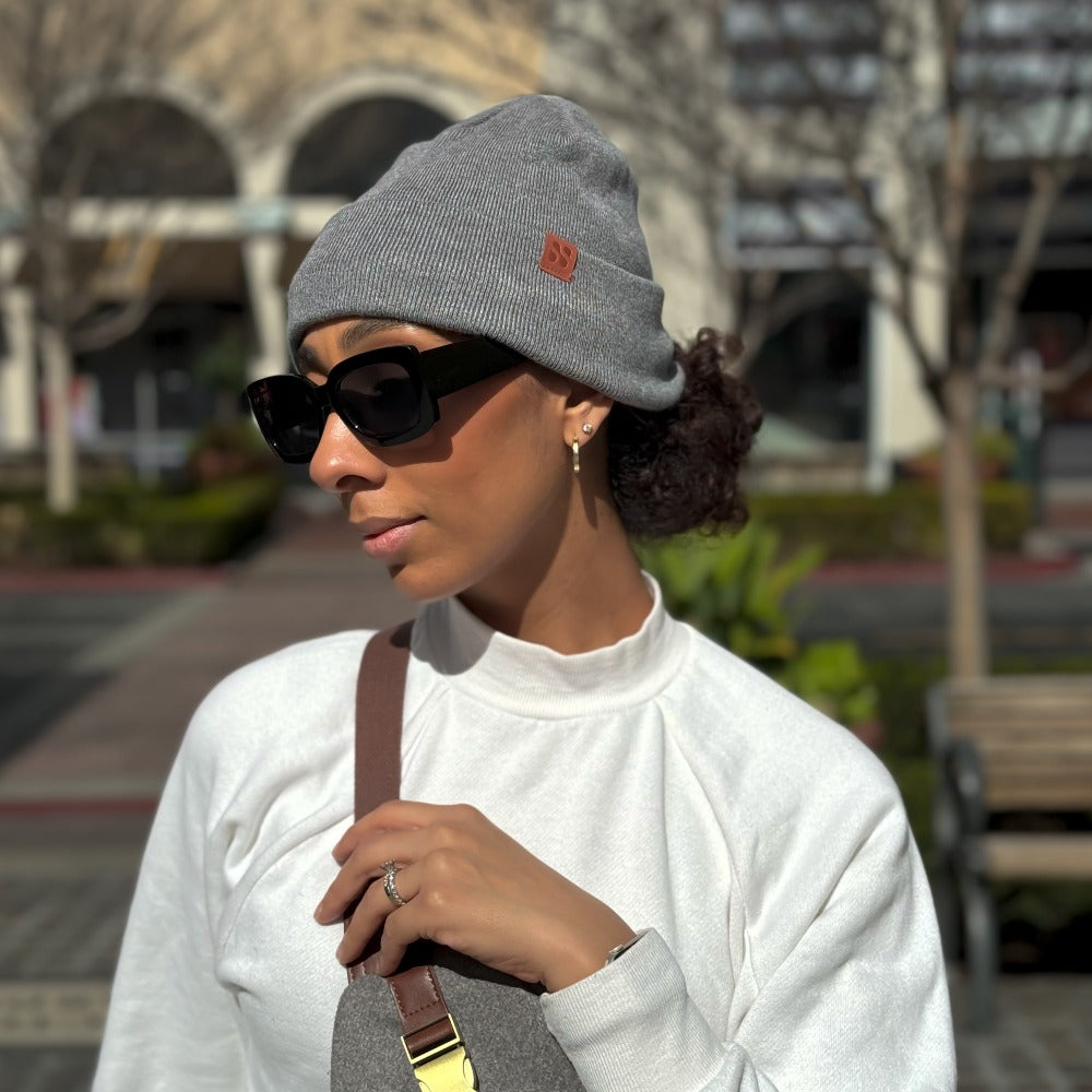 Woman wearing gray SafeSleeve Beanie with sunglasses and white shirt, standing outdoors in urban setting.