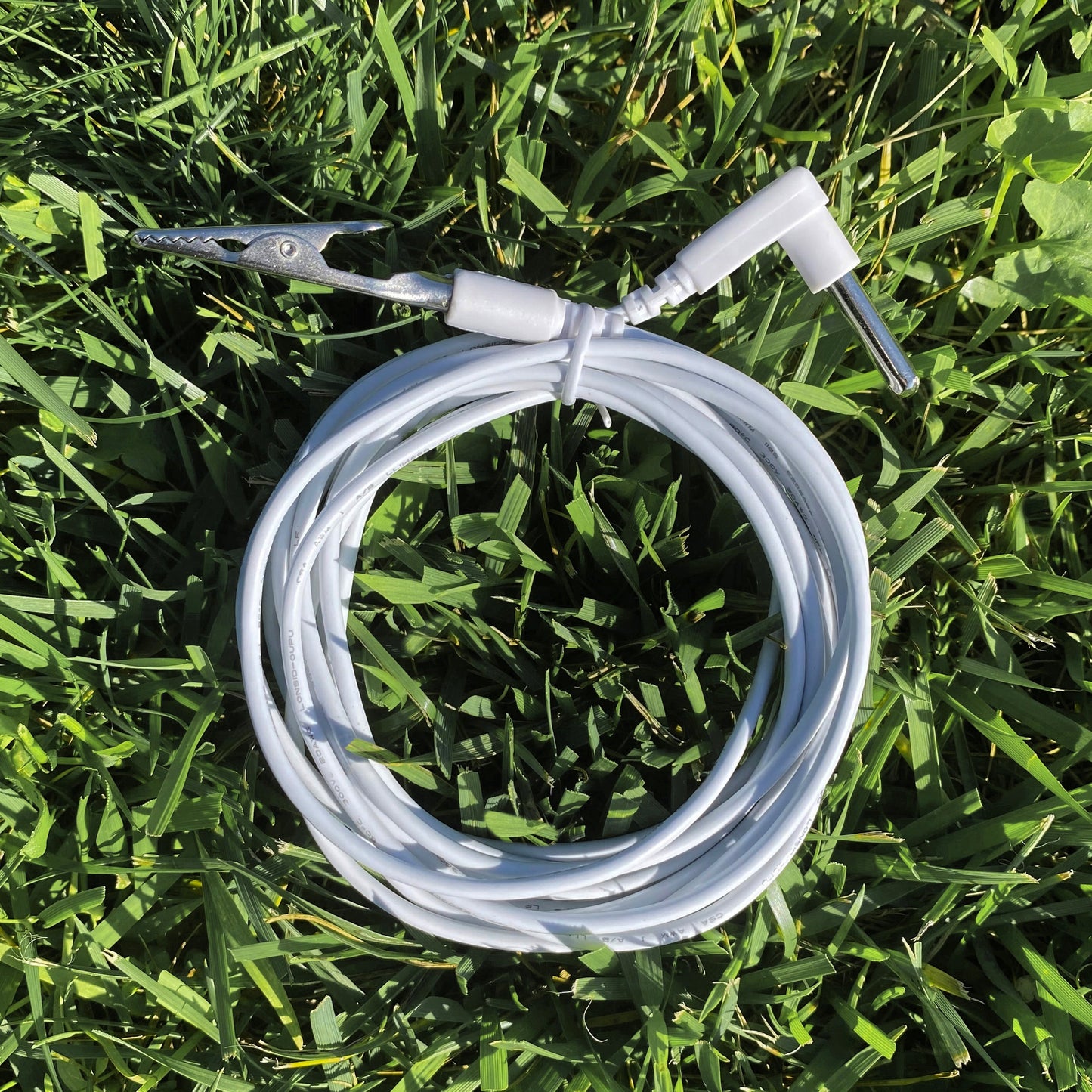 TRU47 15' Grounding Alligator Cord coiled on grass with metal alligator clip and white connectors visible.