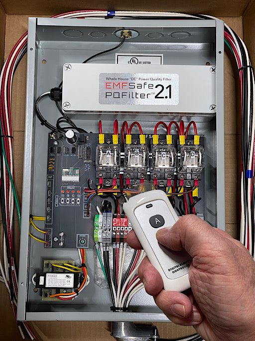 Hand holding white remote control above open EMFSafe PoFilter 2.1 electrical panel with circuit breakers and wiring.
