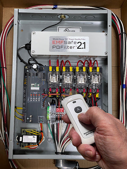 Hand holding white remote control above open EMFSafe PoFilter 2.1 electrical panel with circuit breakers and wiring.