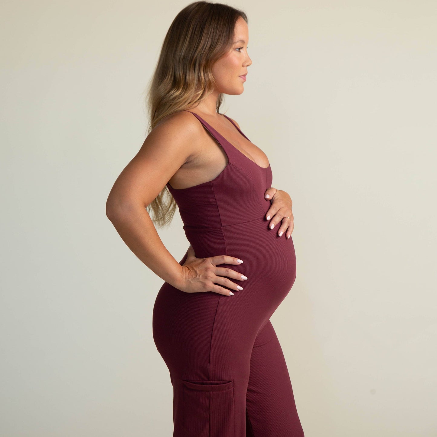 Pregnant woman wearing burgundy Juunaday Everywhere Playsuit in side profile, hands on belly, against neutral background.
