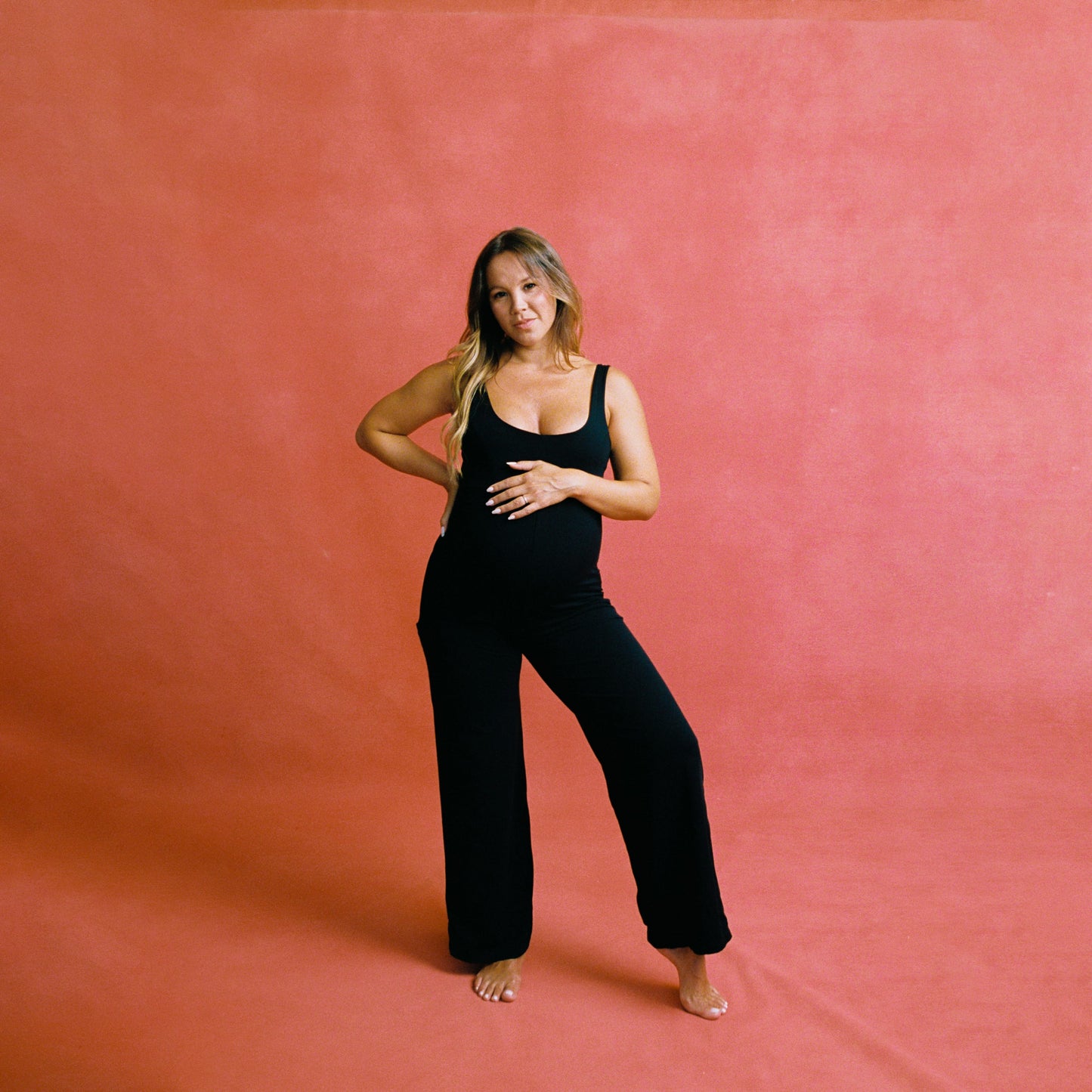 Pregnant woman wearing black Juunaday Everywhere Playsuit with wide legs, standing barefoot against coral gradient background.