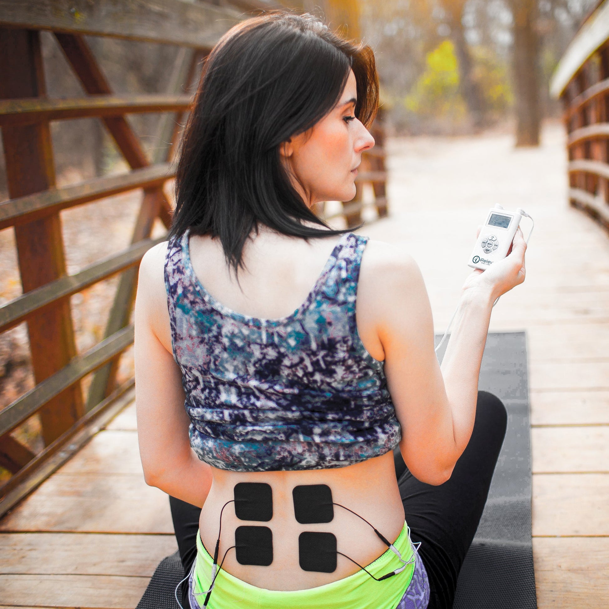 Woman wearing iReliev TENS electrode pads on lower back, holding wireless remote control device outdoors.