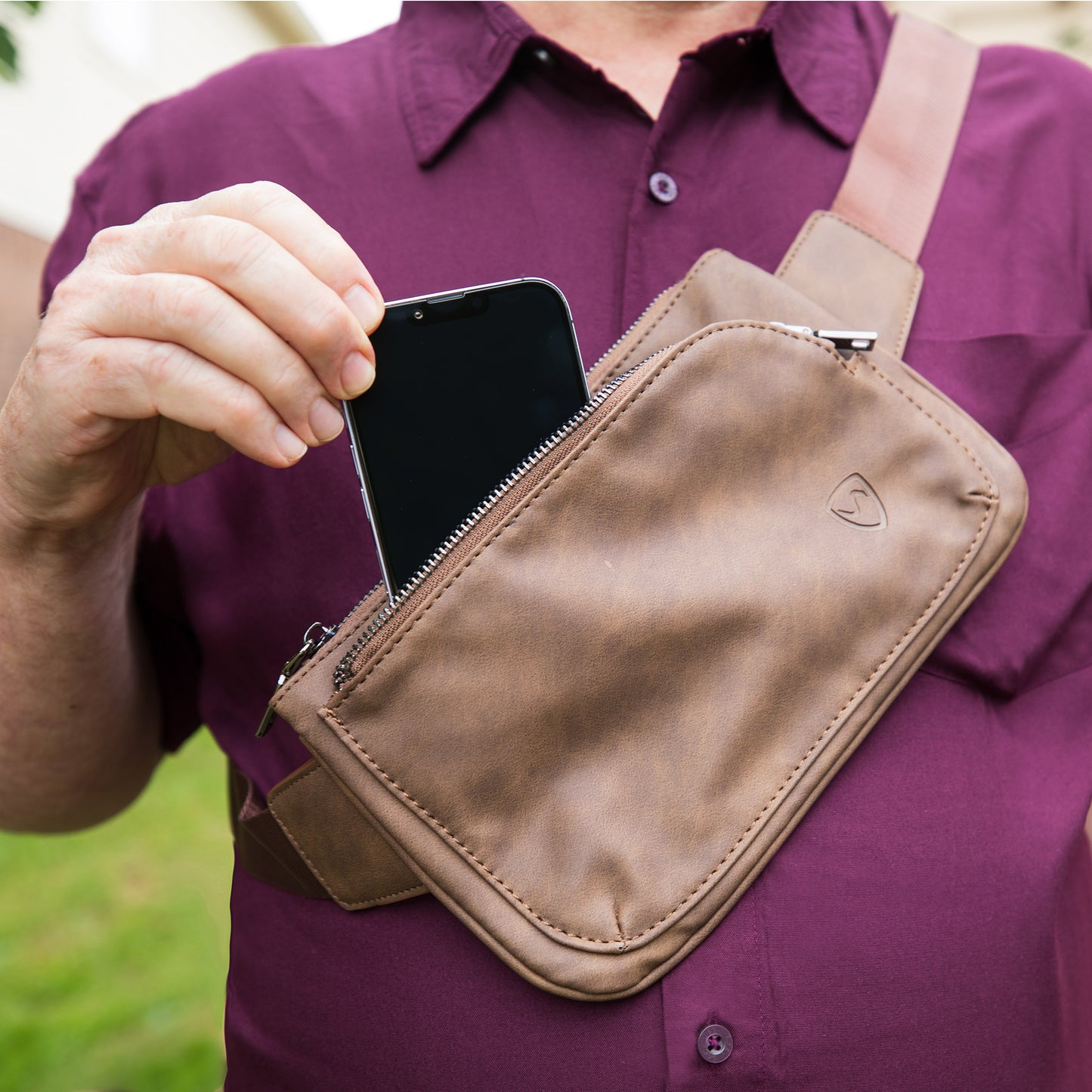Person in purple shirt holding smartphone while wearing tan SYB Cross Body Fanny Pack with heart logo, shown in use outdoors.