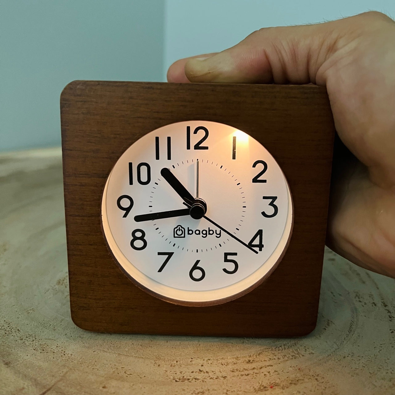 Hand holding Bagby silent analog alarm clock with wood base and white face, displaying 9 o'clock.