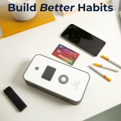 Mindsight Timed Lock Box displayed on white desk with smartphone, credit card, cigarettes, and yellow notebook nearby.