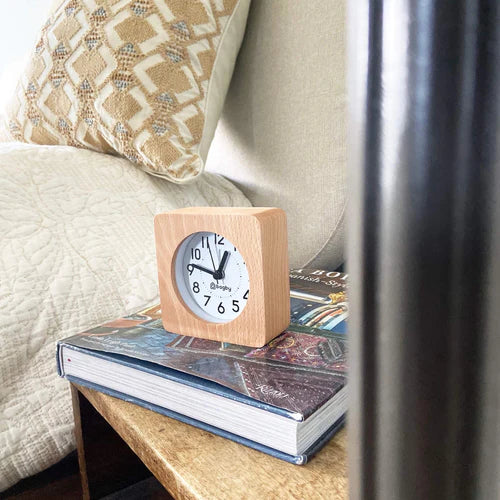Wooden alarm clock displayed on stacked books atop a nightstand next to a bed with pillows and curtains.