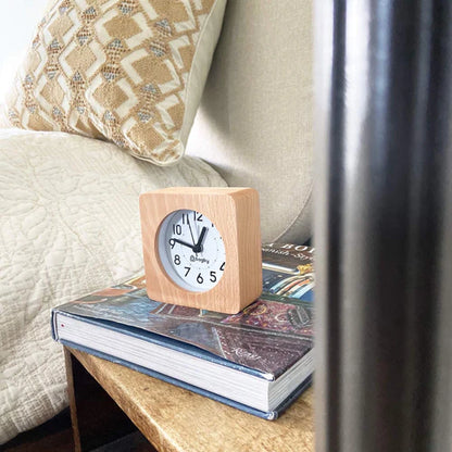 Wooden alarm clock displayed on stacked books atop a nightstand next to a bed with pillows and curtains.